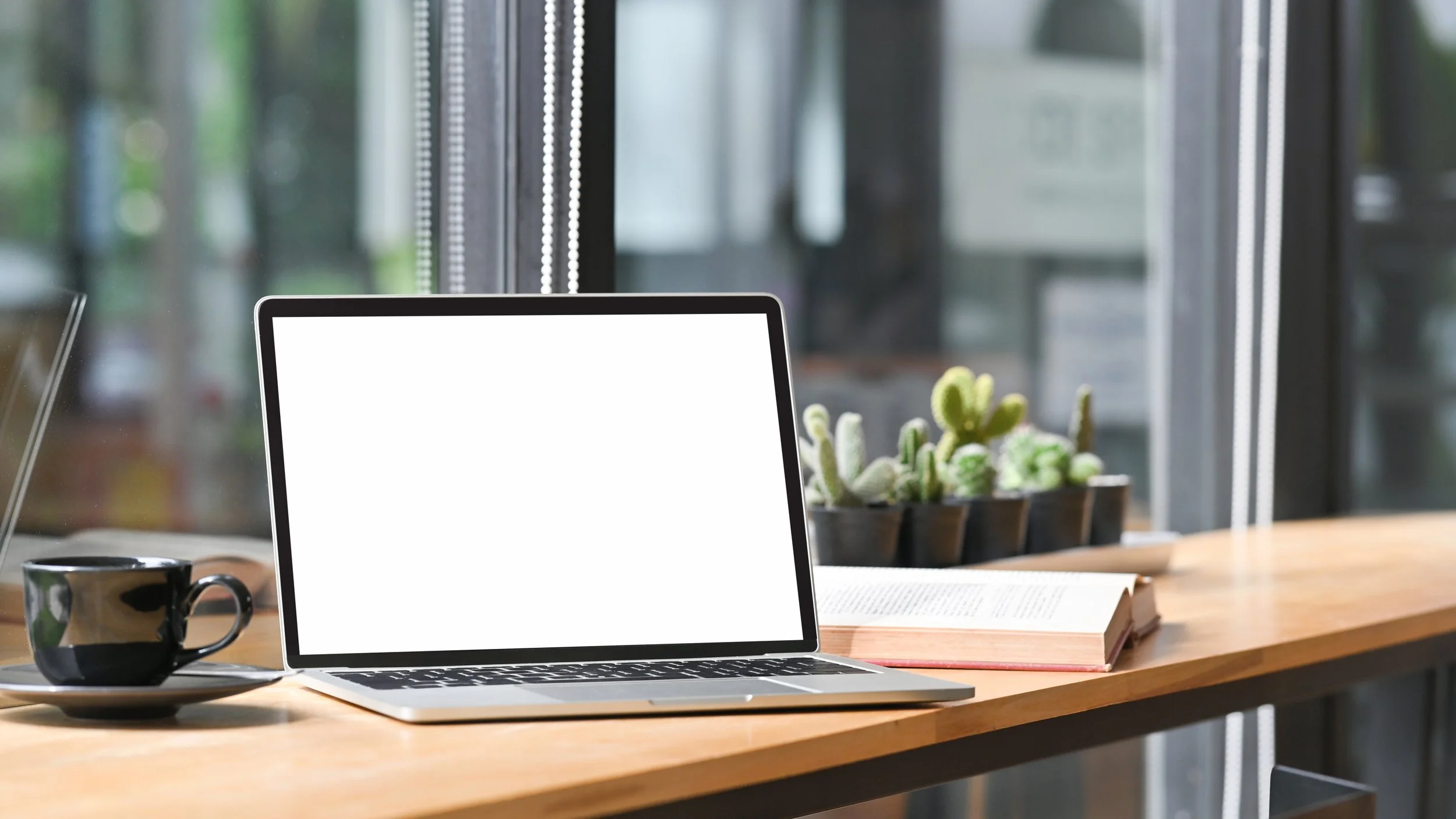A modern workspace with a laptop, black coffee mug, open book, and a rectangular planter with green succulents on a wooden desk near large windows.