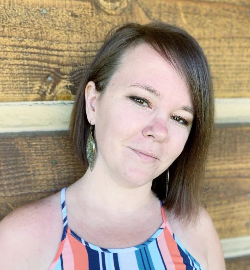 A woman with medium brown hair wearing a colorful striped tank top and large earrings, standing in front of a wooden wall.