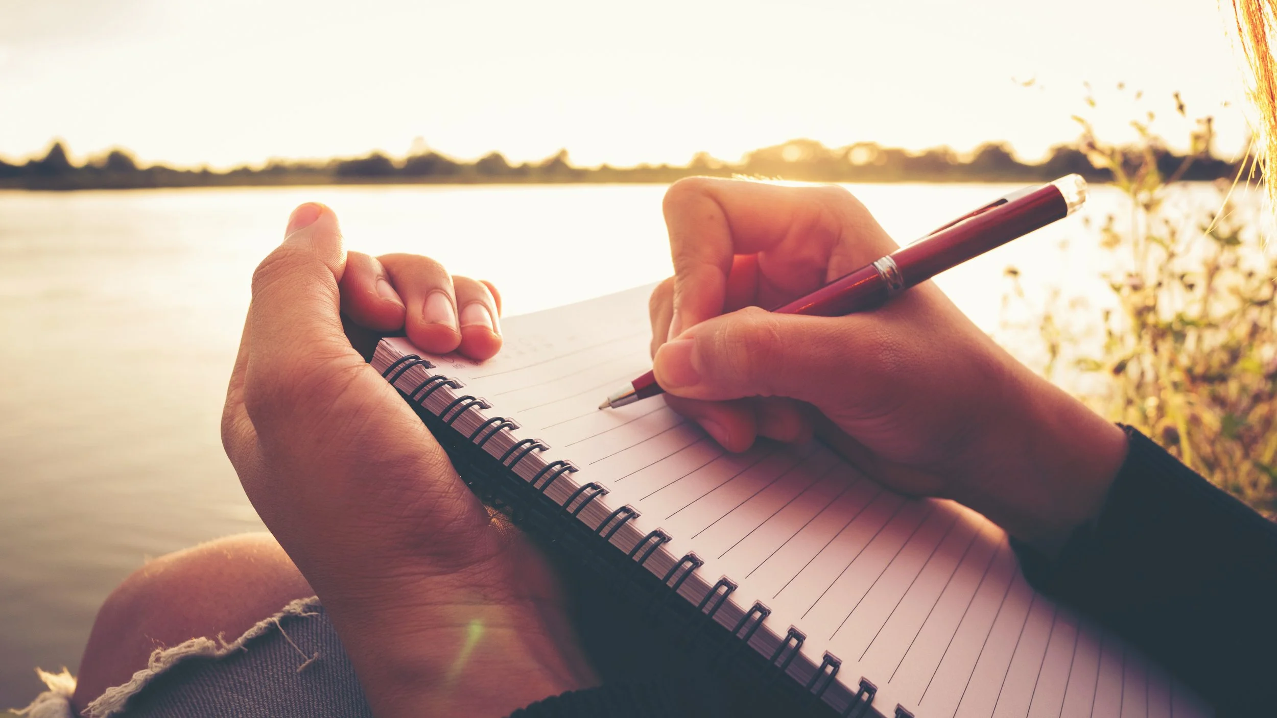 A person holding a spiral notebook and pen near a body of water at sunset, with sunlight reflecting on the water and plants nearby.