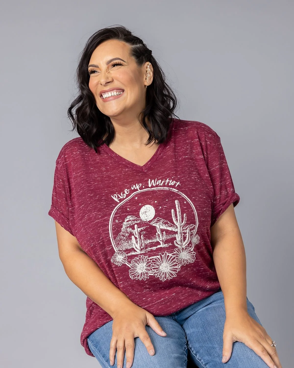 A woman with dark wavy hair, wearing a maroon T-shirt with white desert-themed graphics and the words "Rise up Warrior," sitting against a gray background, smiling and squinting.