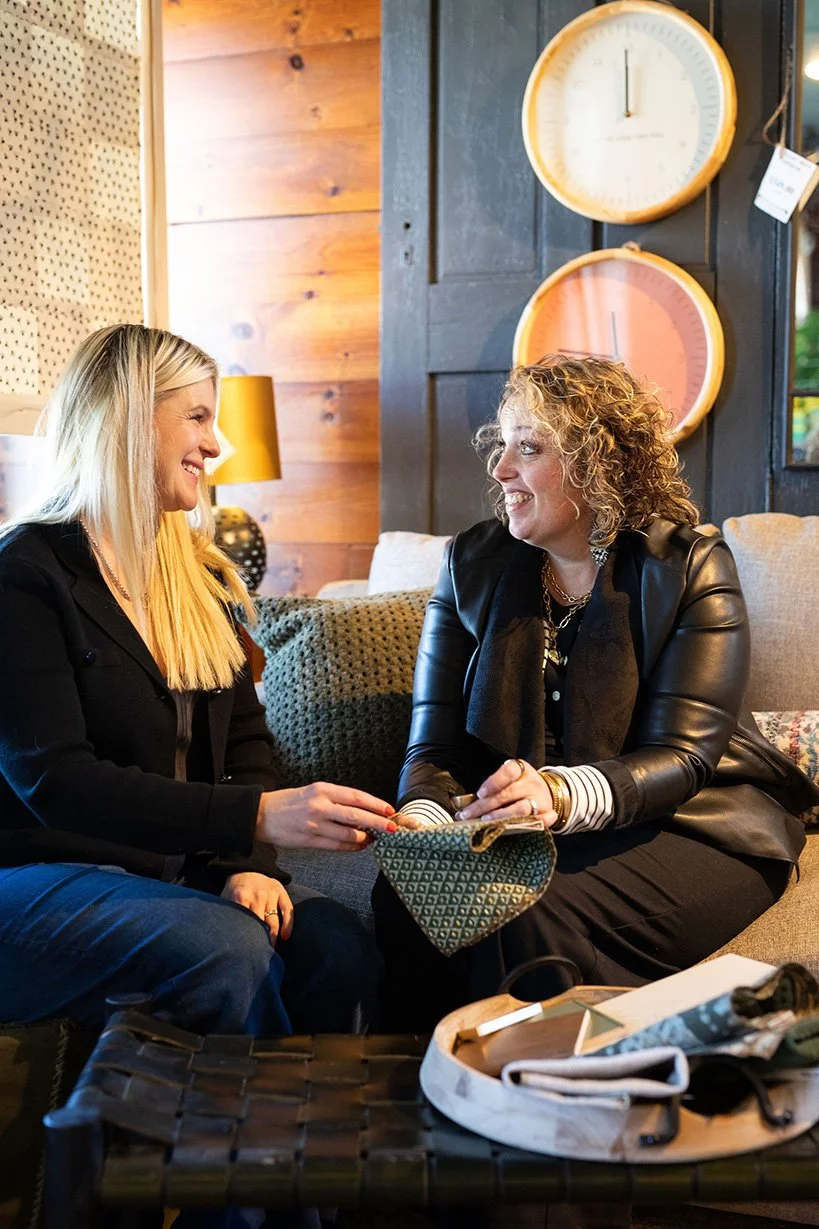Two women sitting on a gray couch, smiling and talking to each other in a cozy room with wooden walls and large clocks on the wall.