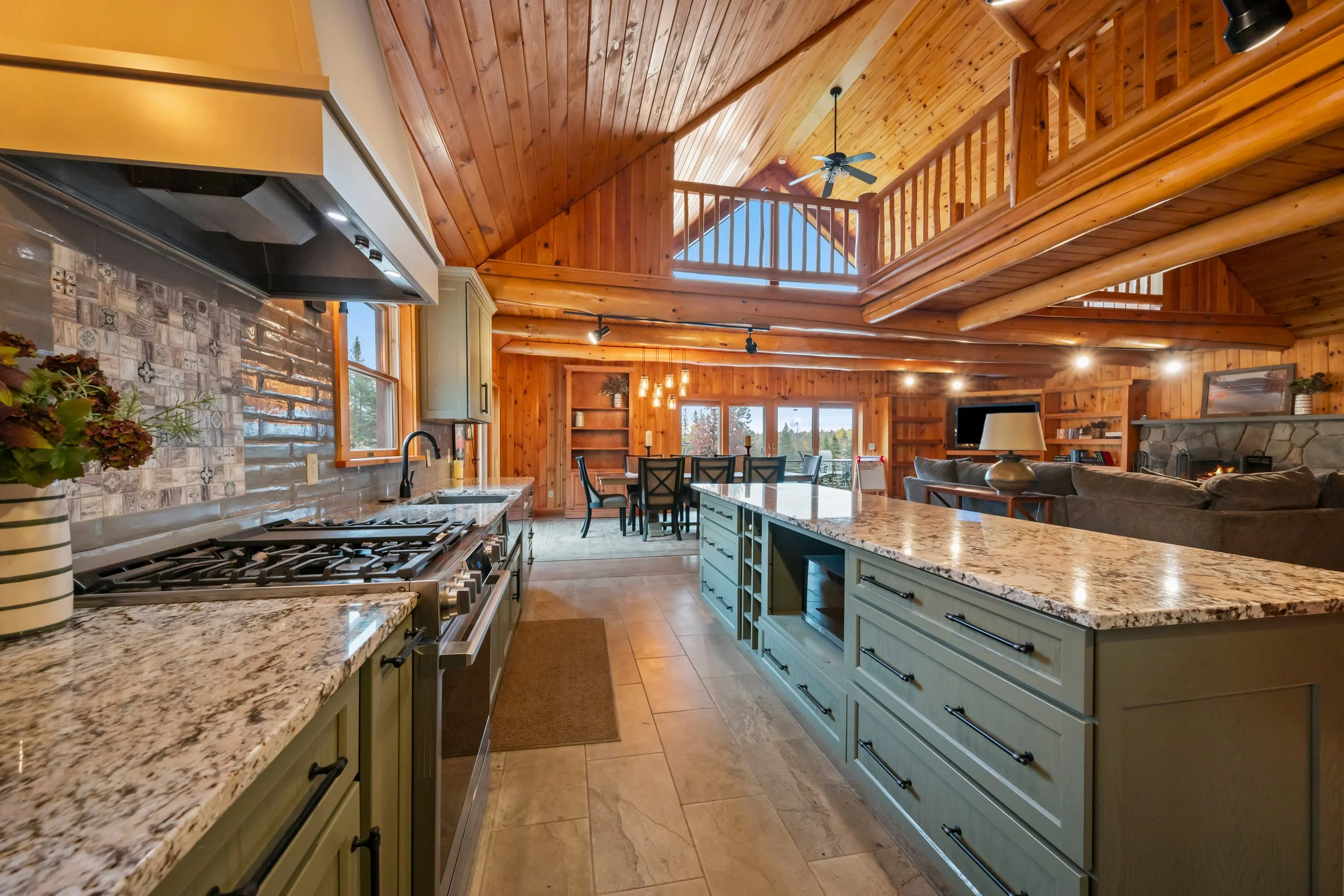 Open kitchen and living area with wooden walls and ceiling, granite countertops, stove, dining table, and seating area with TV and fireplace.