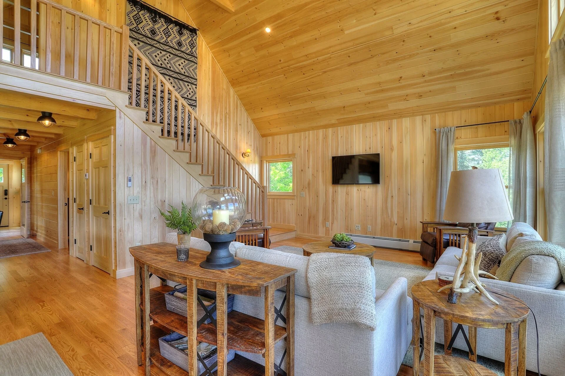 Living room with wood-paneled walls and ceiling, featuring a sofa, side tables, a wall-mounted TV, windows with curtains, a staircase with wooden railing, and hardwood floors.