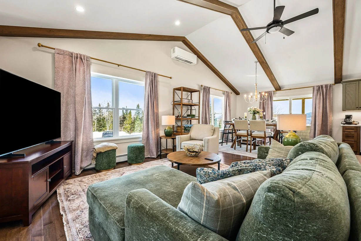 A bright, spacious living room with large windows, a green velvet sofa, a beige armchair, a wooden coffee table, and a flat-screen TV on a wooden console. There are curtains, a ceiling fan, a chandelier, and a dining area with a table and chairs in the background.