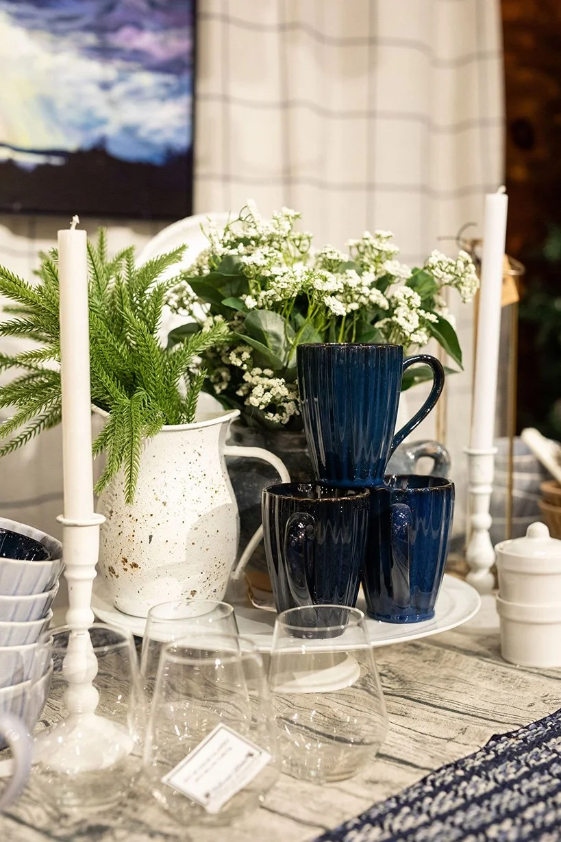 Decorative table setting at the North & Found shop, with white and blue cups, clear glasses, white candlesticks, potted greenery, and a flower arrangement on a rustic wooden surface.