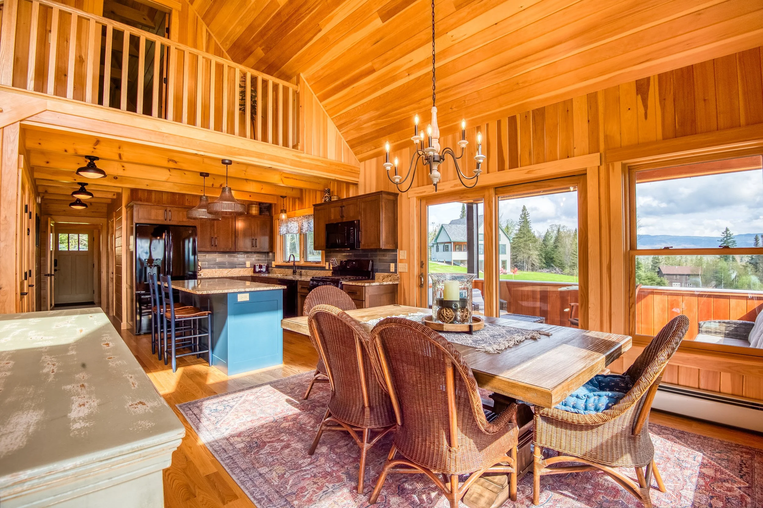 Interior of a cozy wooden cabin with a dining area, a kitchen with dark cabinets and an island, a chandelier, and large windows showcasing outdoor scenery.