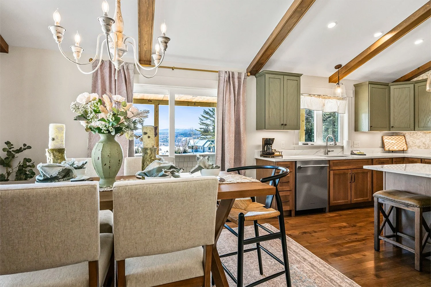 A cozy kitchen and dining area with a wooden table, beige chairs, a floral centerpiece, and a view of a snowy landscape through a sliding glass door.