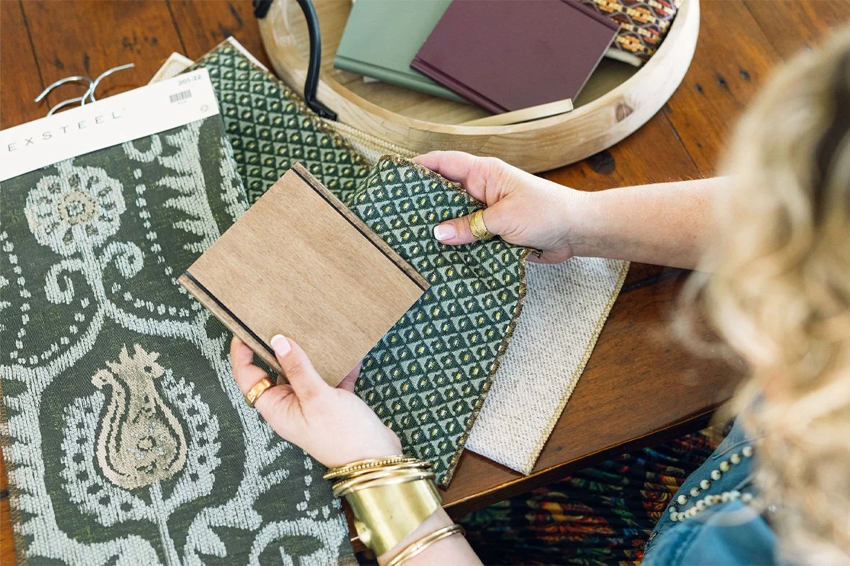 Virginia Patnode MacFawn holding fabric samples and a small wood sample, sitting at a table with fabric swatches, color samples, and a tray of fabric samples.
