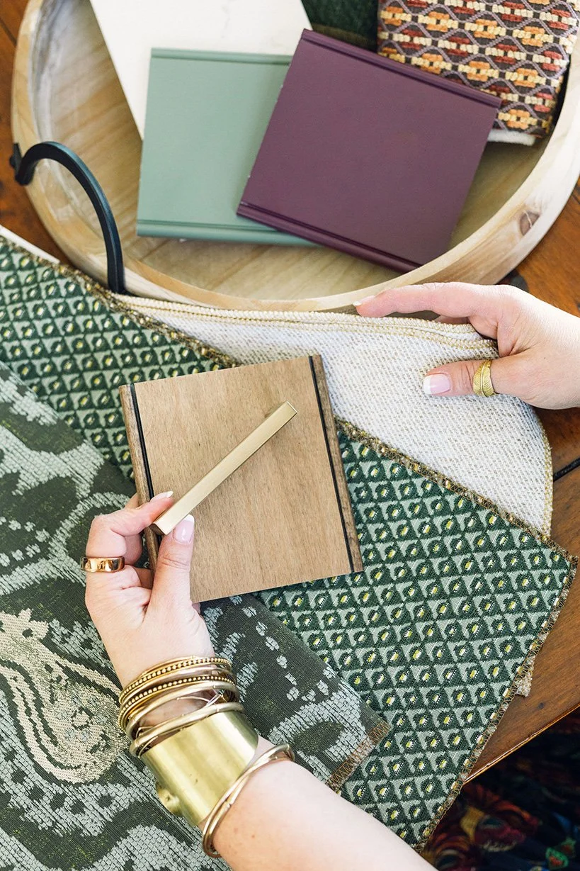 Close-up of Virginia Patnode MacFawn holding a small wooden sample board with a beige handle, sitting at a table with fabric samples, paint chips, and a plate of color swatches.