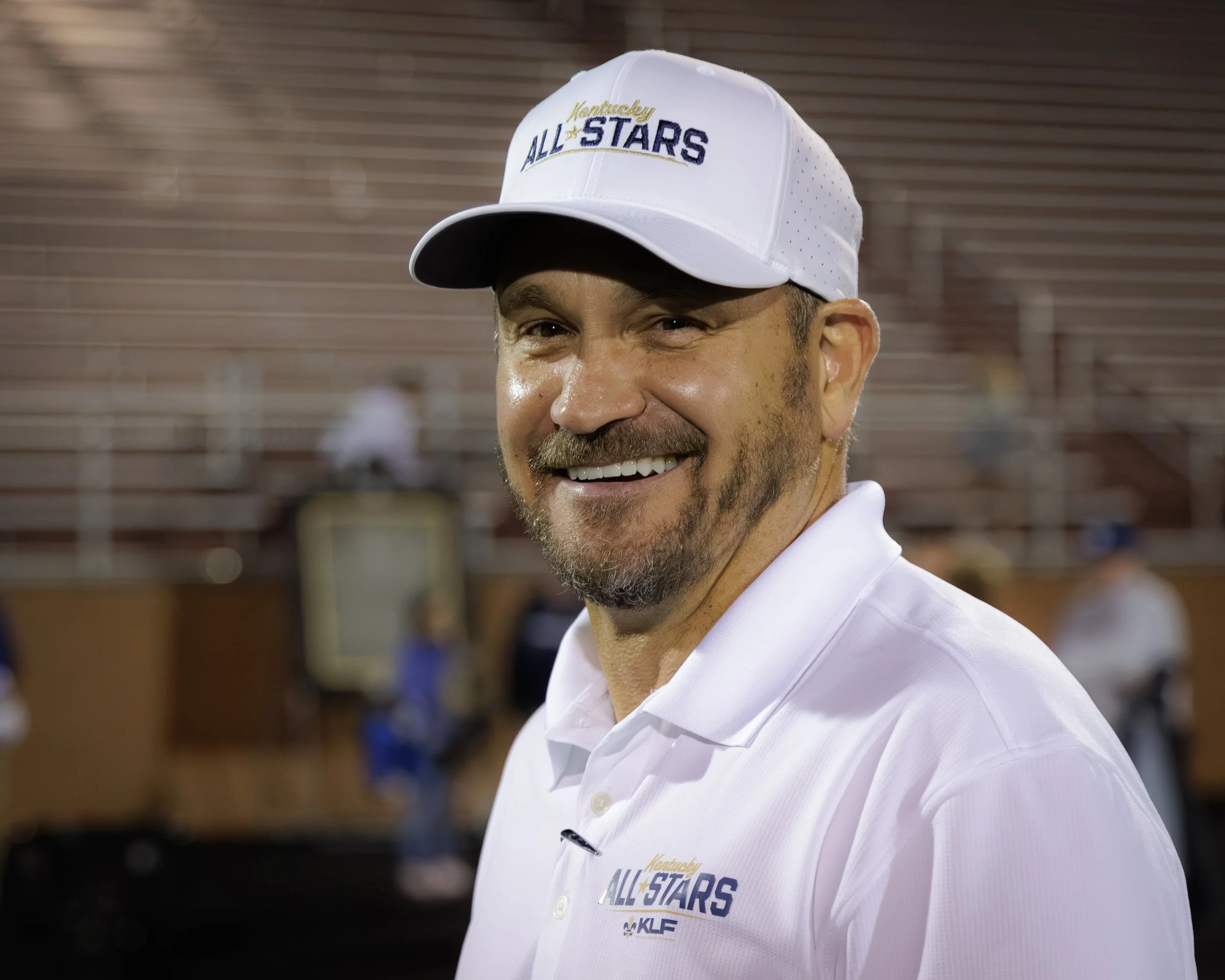 A smiling man wearing a white hat and shirt with "Kentucky All Stars" logos, standing in front of stadium seating.