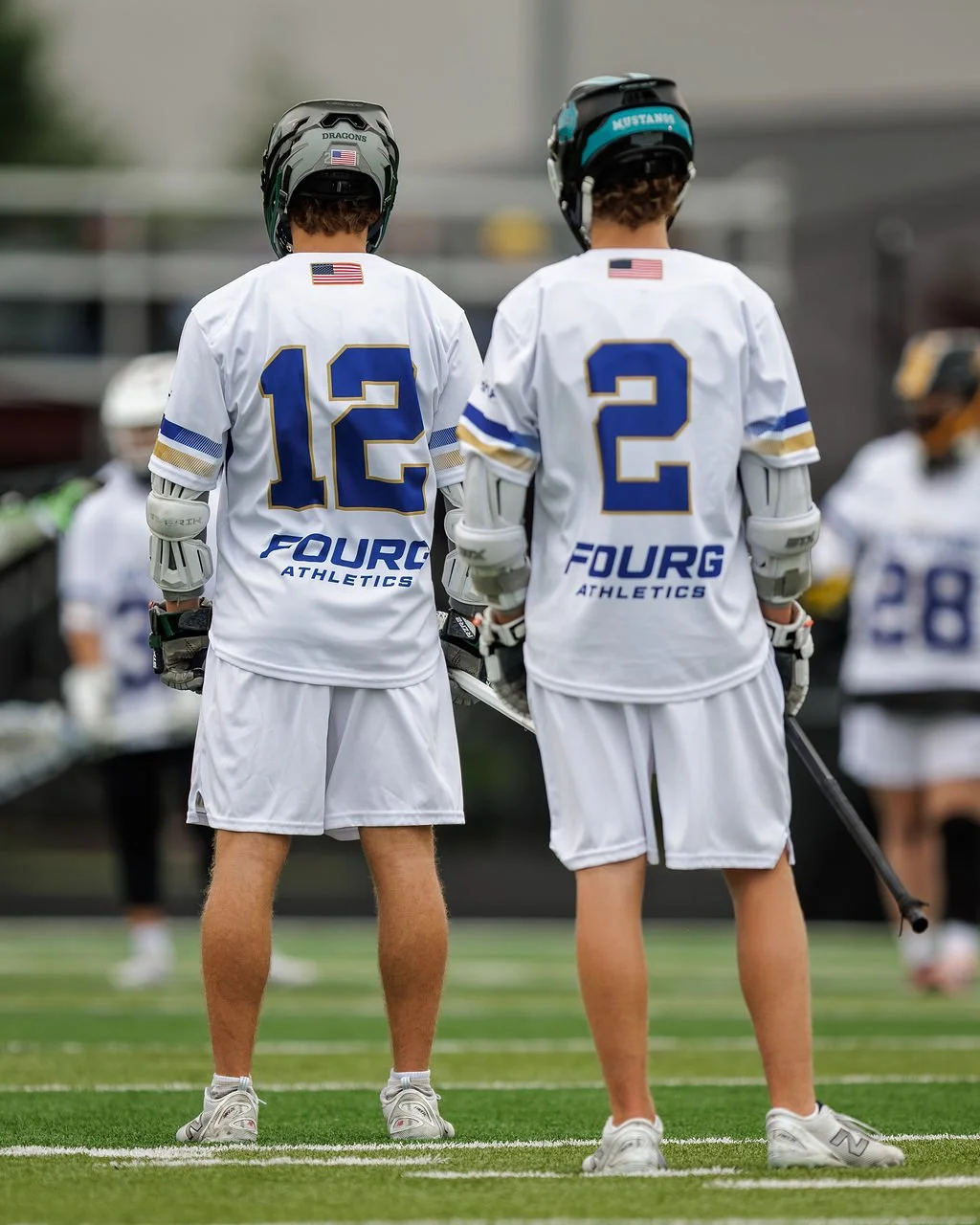 Two lacrosse players stand on a field with their backs to the camera, wearing white jerseys with blue and gold accents, helmets, and holding lacrosse sticks.
