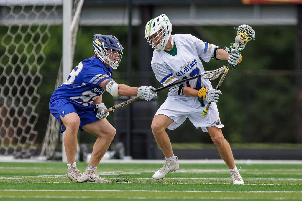 Two lacrosse players competing for the ball during a game on a green field, one wearing a white uniform and the other in blue, both with protective helmets and gloves.