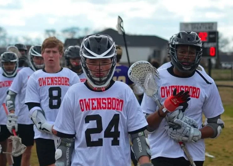 Lacrosse players from Owensboro team in white jerseys with red lettering, wearing helmets and holding lacrosse sticks, walking on a field.