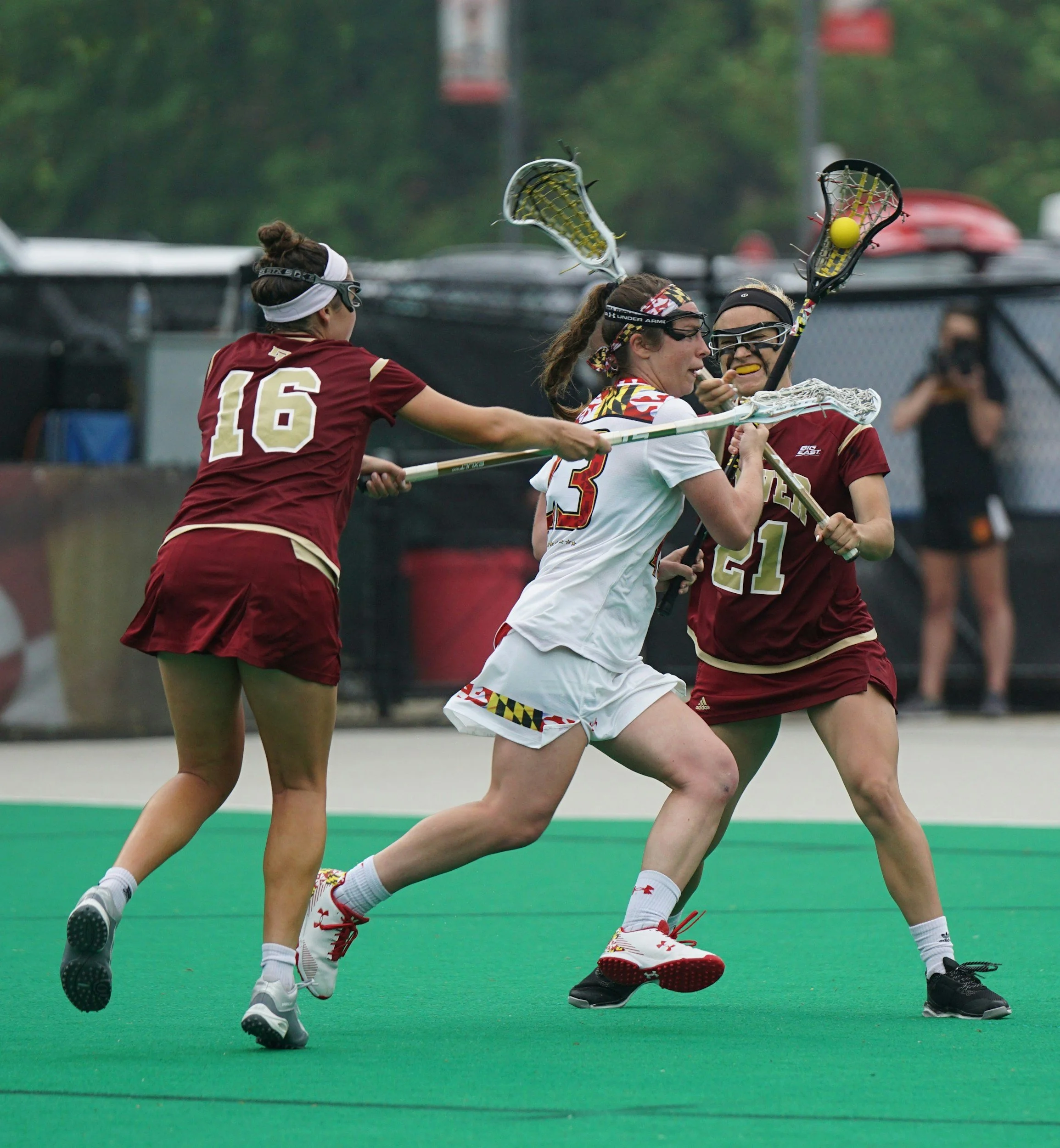 Lacrosse players in maroon and white uniforms competing for possession of the ball on a green field.