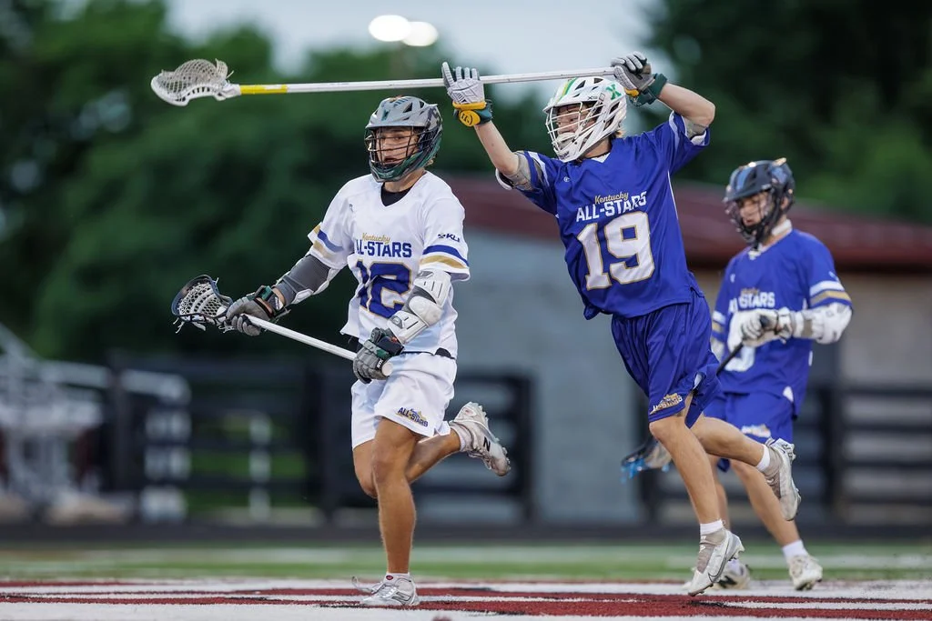 Lacrosse players competing during a game, with one player in a white jersey and two players in blue jerseys, all wearing helmets and protective gear.