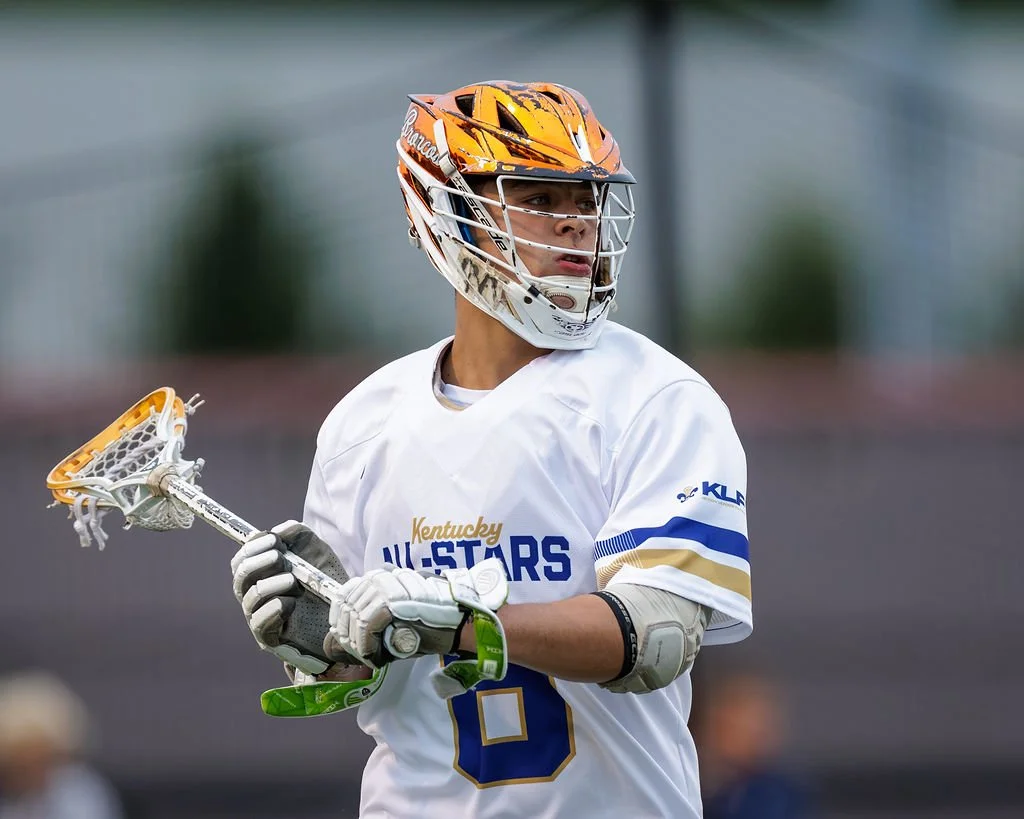 A lacrosse player wearing a helmet and white jersey with blue and gold accents, holding a lacrosse stick on the field.