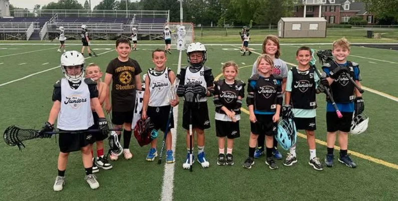 Group of young boys and girls in lacrosse uniforms posing together on a field with teammates in the background.