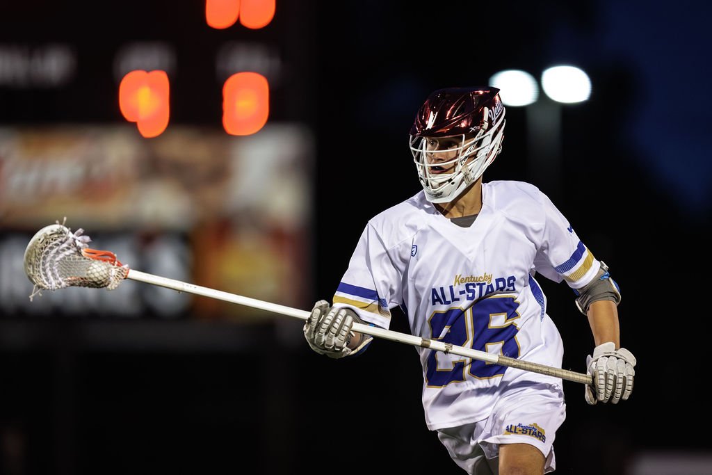 Lacrosse player in white jersey and helmet holding a lacrosse stick during a game.