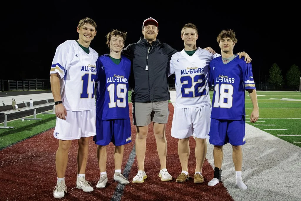 Group of four young male football players and one adult male coach standing on a football field at night, smiling for a photo, wearing jerseys with 'Kentucky All-Stars' printed on them.
