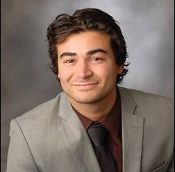 Portrait of a young man with dark hair, wearing a light gray suit and dark shirt, smiling against a gray background.