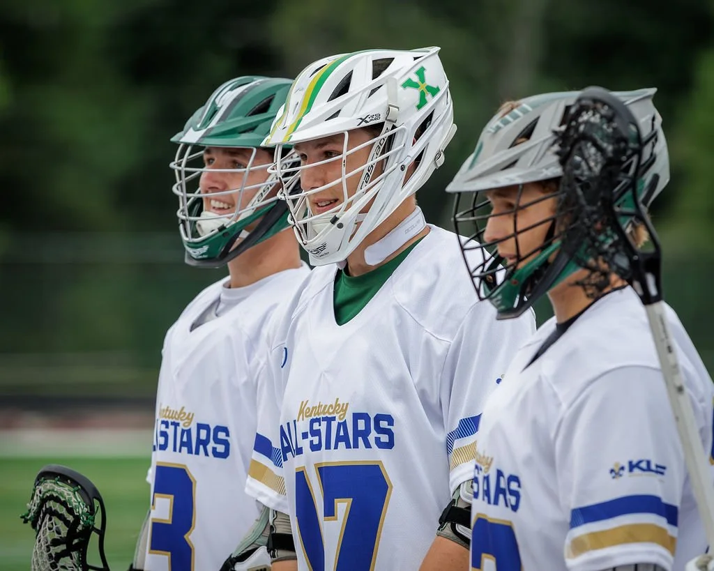 Three male lacrosse players in white jerseys standing in a line, wearing helmets and holding lacrosse sticks, outdoors on a field with trees in the background.