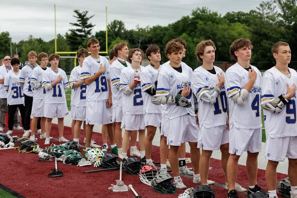 Lacrosse team standing in line on a field during the national anthem, with helmets and gloves on the ground in front of them.