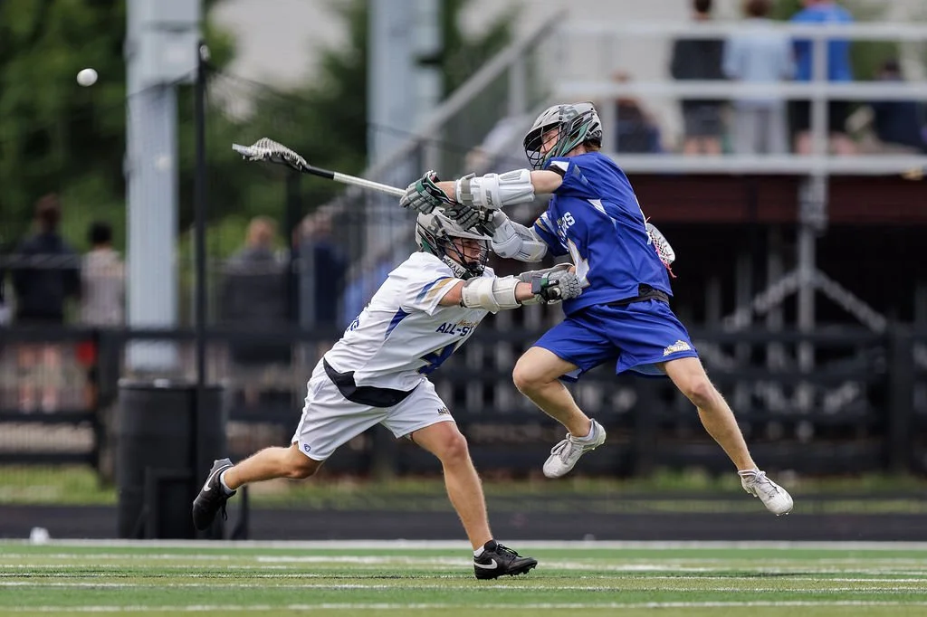 Two young boys in sports uniforms and helmets are playing lacrosse on a field. One boy is jumping, while the other is reaching forward with his lacrosse stick.