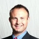 A man with short light brown hair, wearing a dark suit, white shirt, and red tie, smiling against a light background.