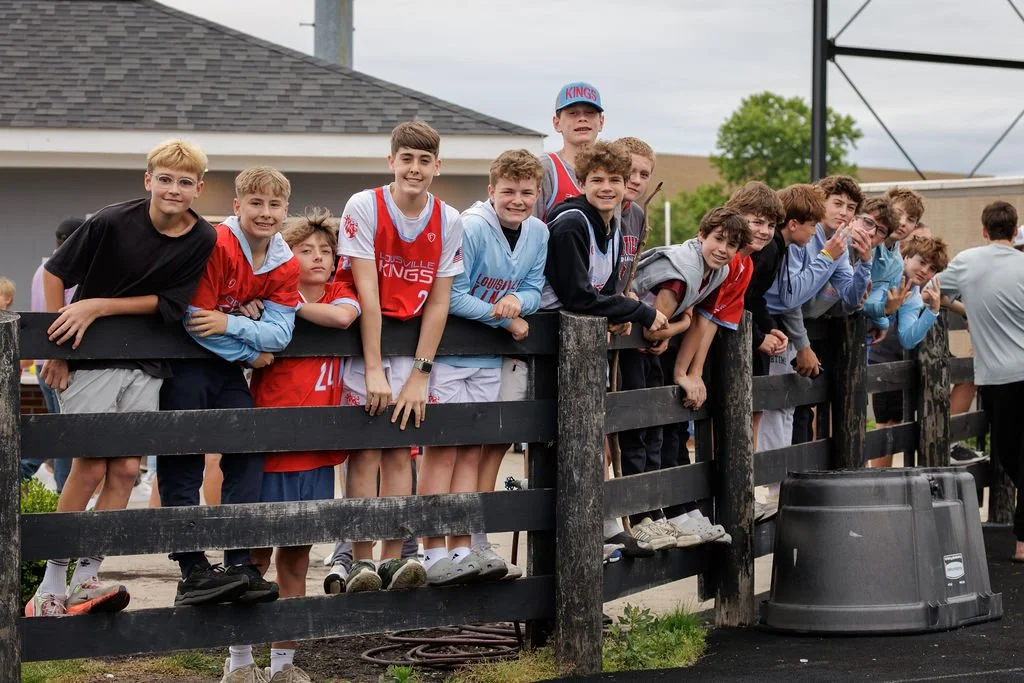 A group of young boys leaning on a black wooden fence at a sports event, some wearing sports jerseys and others in casual clothes, with a building and cloudy sky in the background.