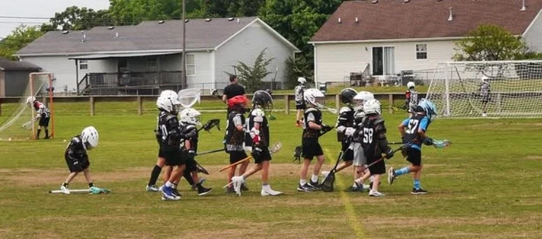 Group of young children playing lacrosse on a grassy field, with some wearing helmets and sports gear, while a coach or adult supervises in the background.