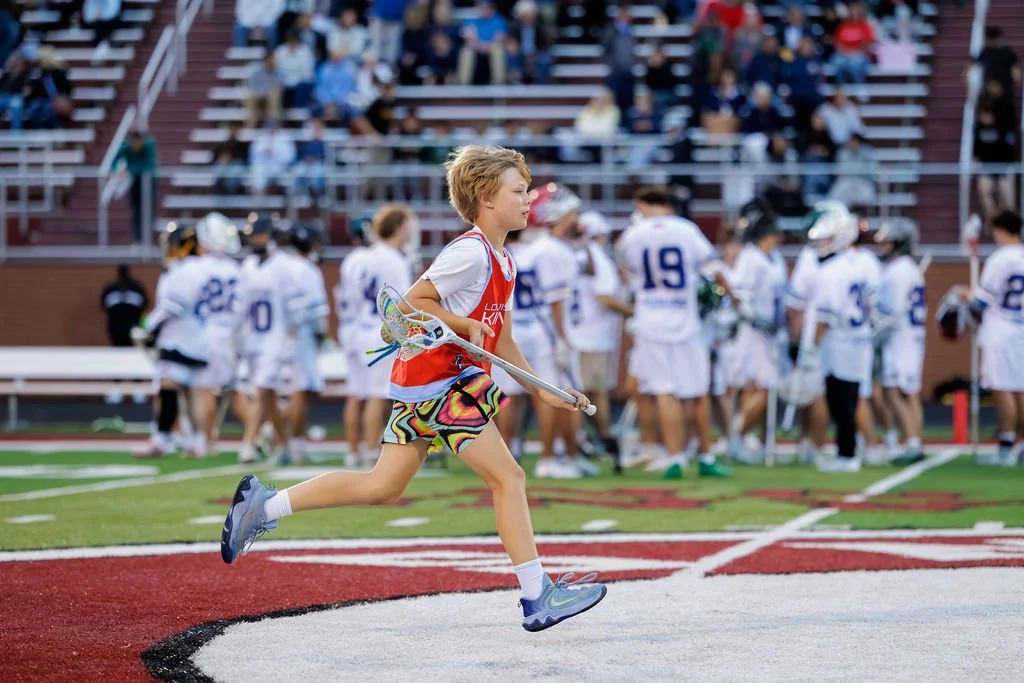 Young girl running on a sports field with a lacrosse stick, with a group of people in sports uniforms and spectators seated in the background.