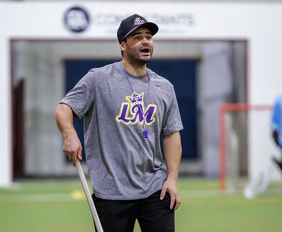 A man in sports attire standing on an indoor field, wearing a gray T-shirt with LUM logo, a baseball cap, and a whistle around his neck, appearing to give instructions or coaching.