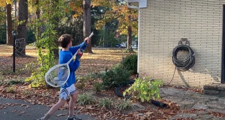 A boy in a blue shirt and striped shorts using a garden hose to spray water on a house exterior in a backyard with trees and fallen leaves.