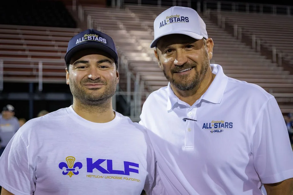 Two men smiling at a sports event, wearing white and navy hats and shirts with 'Kentucky All Stars' and 'KLF' logos, standing in a stadium with empty bleachers in the background.