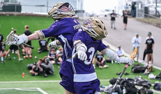 Two lacrosse players in purple uniforms celebrating on a field, with fans and others in the background.