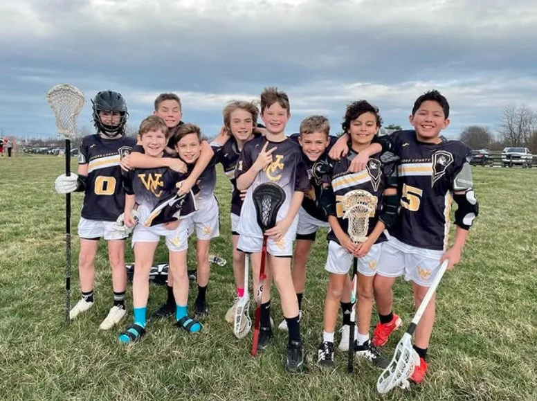 Group of young boys playing lacrosse on a grassy field, wearing sports jerseys and shorts, holding lacrosse sticks, smiling after a game.