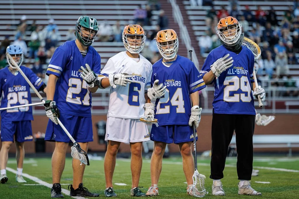 Lacrosse players in uniforms with helmets and gear on a field at a sports event.