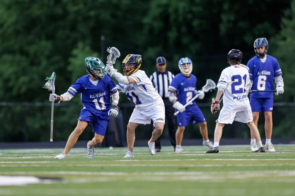 Children playing lacrosse on a field, wearing helmets and athletic uniforms, with a referee in the background.