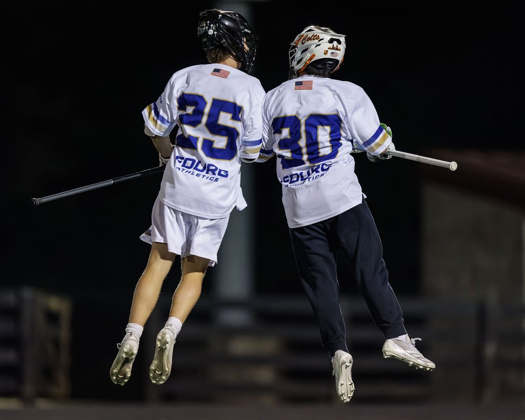 Two athletes in hockey jerseys jumping in the air at night, holding hockey sticks. One wears jersey number 25, the other 30.