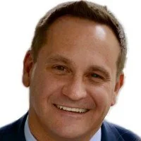 Close-up photo of a smiling man with short brown hair, wearing a suit and tie.
