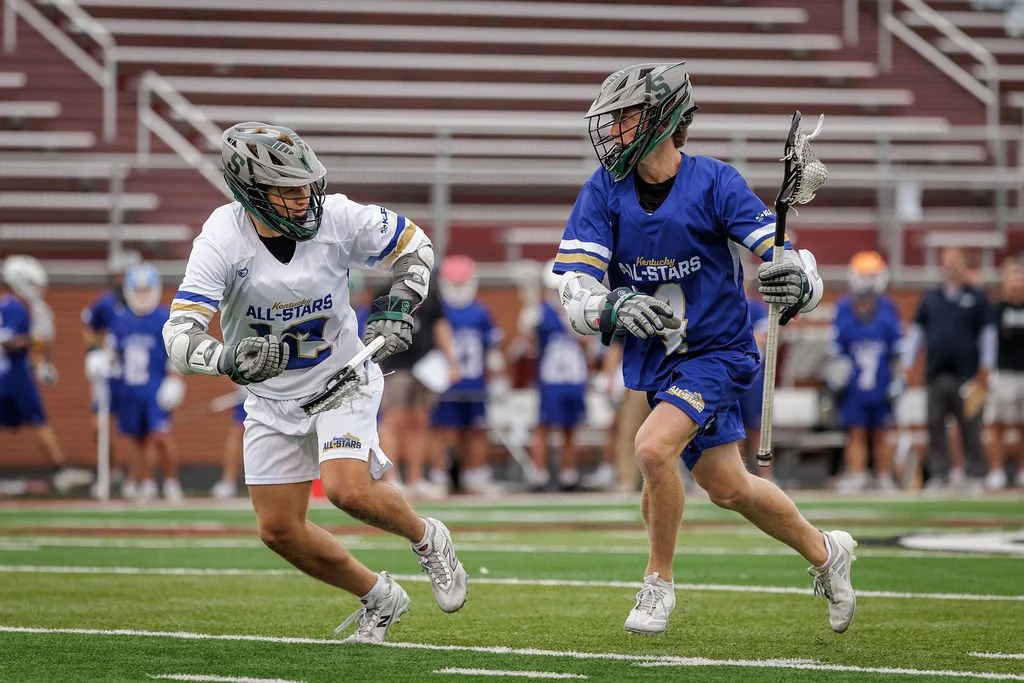 Two lacrosse players in blue and white uniforms playing on the field with spectators in the background.