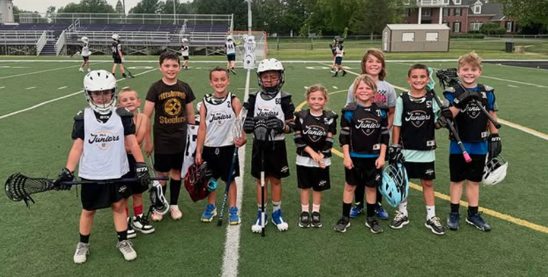 Group of young boys and girls in lacrosse uniforms on a field, holding lacrosse sticks, with other players practicing in the background.