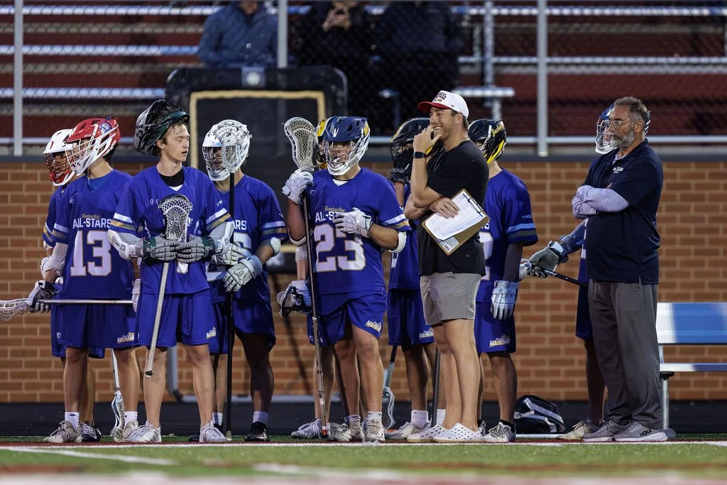 Lacrosse players and coaches on the sideline during a game, with players wearing blue uniforms and helmets, some holding lacrosse sticks, and coaches talking to them.