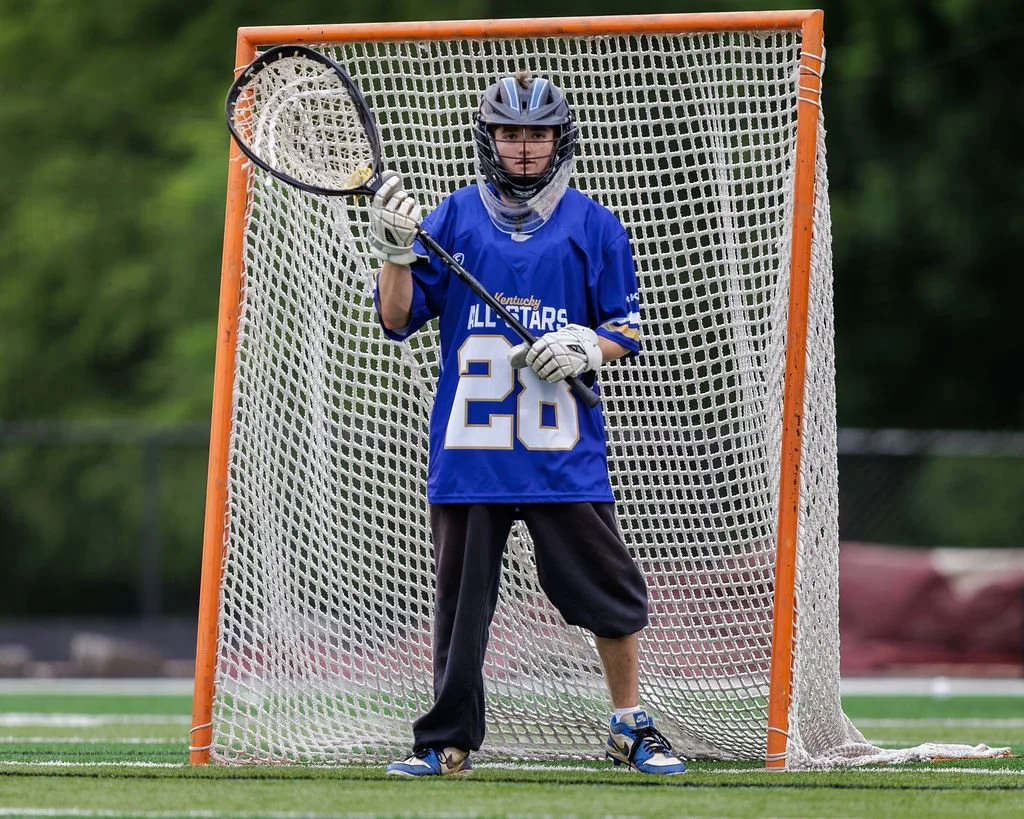 A young male lacrosse goalie standing in front of the goal net on a field, wearing a blue jersey, helmet, and gloves, holding a lacrosse stick.