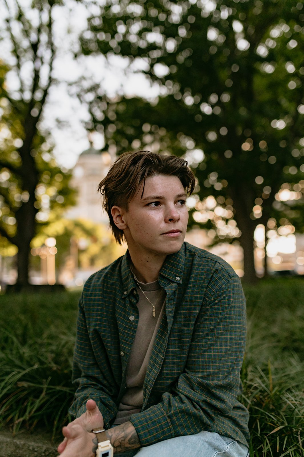 A young person with short brown hair sitting outdoors in front of trees and a building, wearing a green plaid shirt over a beige t-shirt, with accessories including a necklace, a watch, and rings.