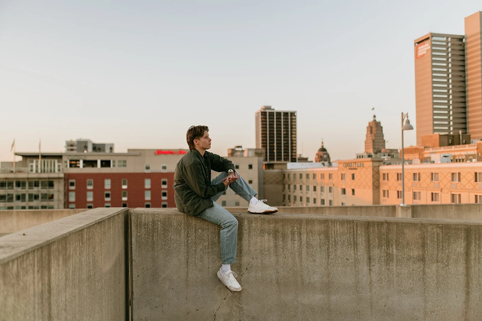A young man with dark hair sitting on a concrete ledge on a rooftop during sunset, with a city skyline featuring various buildings in the background.