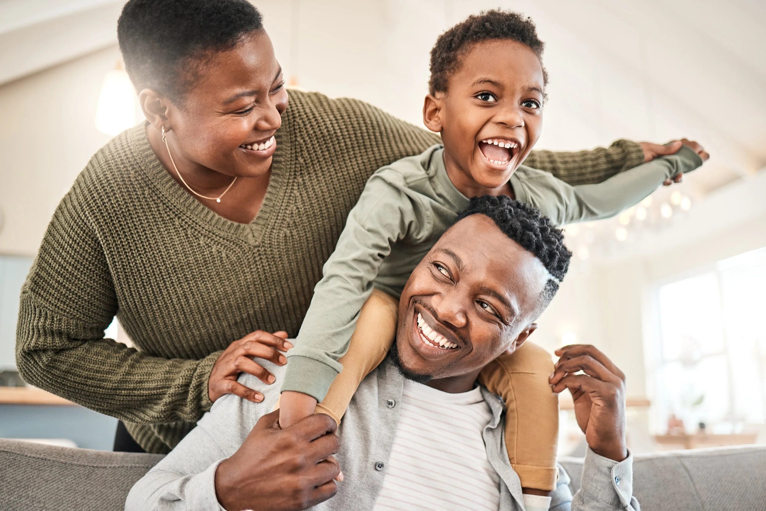 A diverse family enjoying a playful moment together indoors. The family members are smiling and laughing, with a woman and a man holding a young boy on their shoulders.
