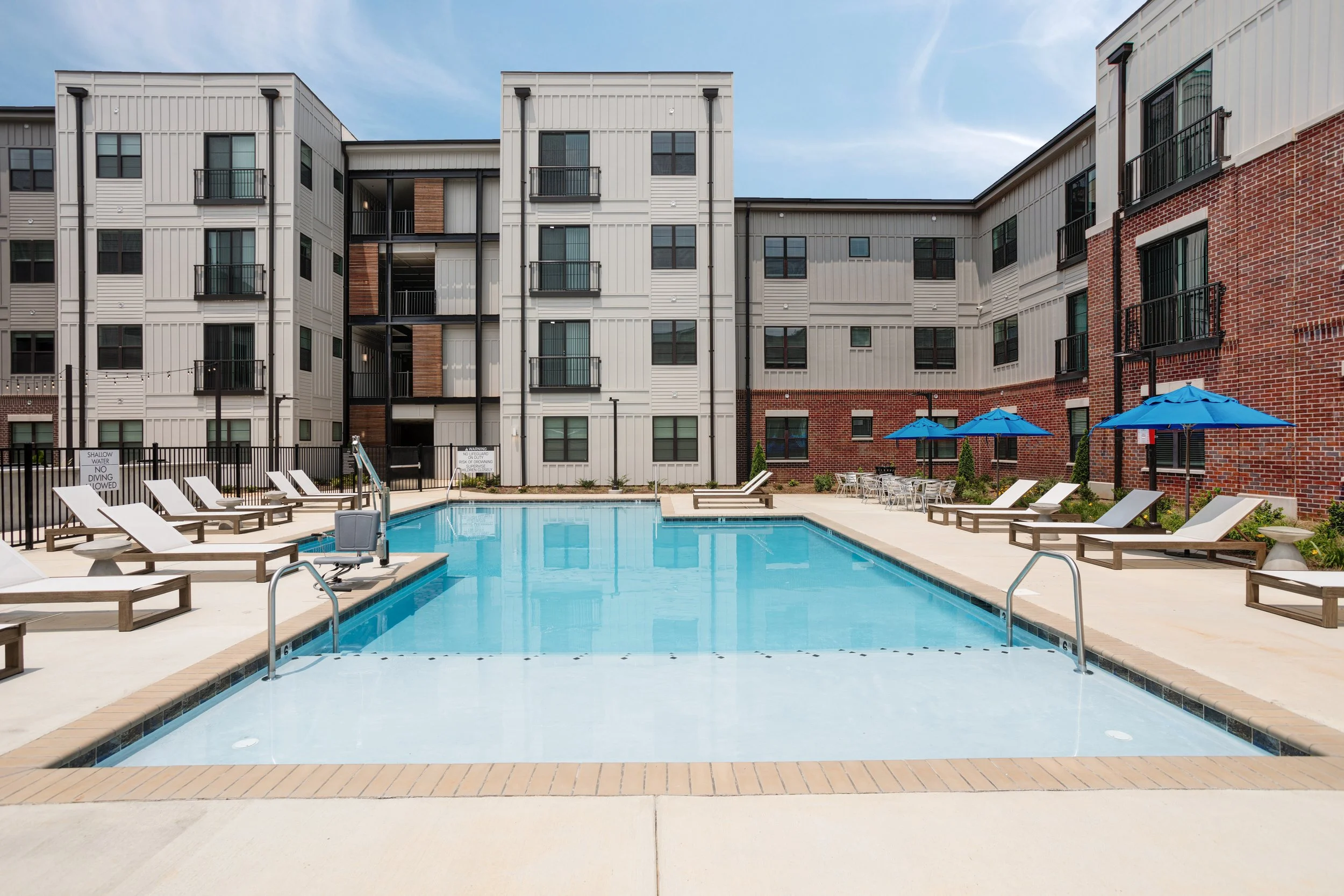 Apartment complex with a swimming pool, lounge chairs, blue umbrellas, and multi-story buildings with balconies and a mix of siding and brick exterior.