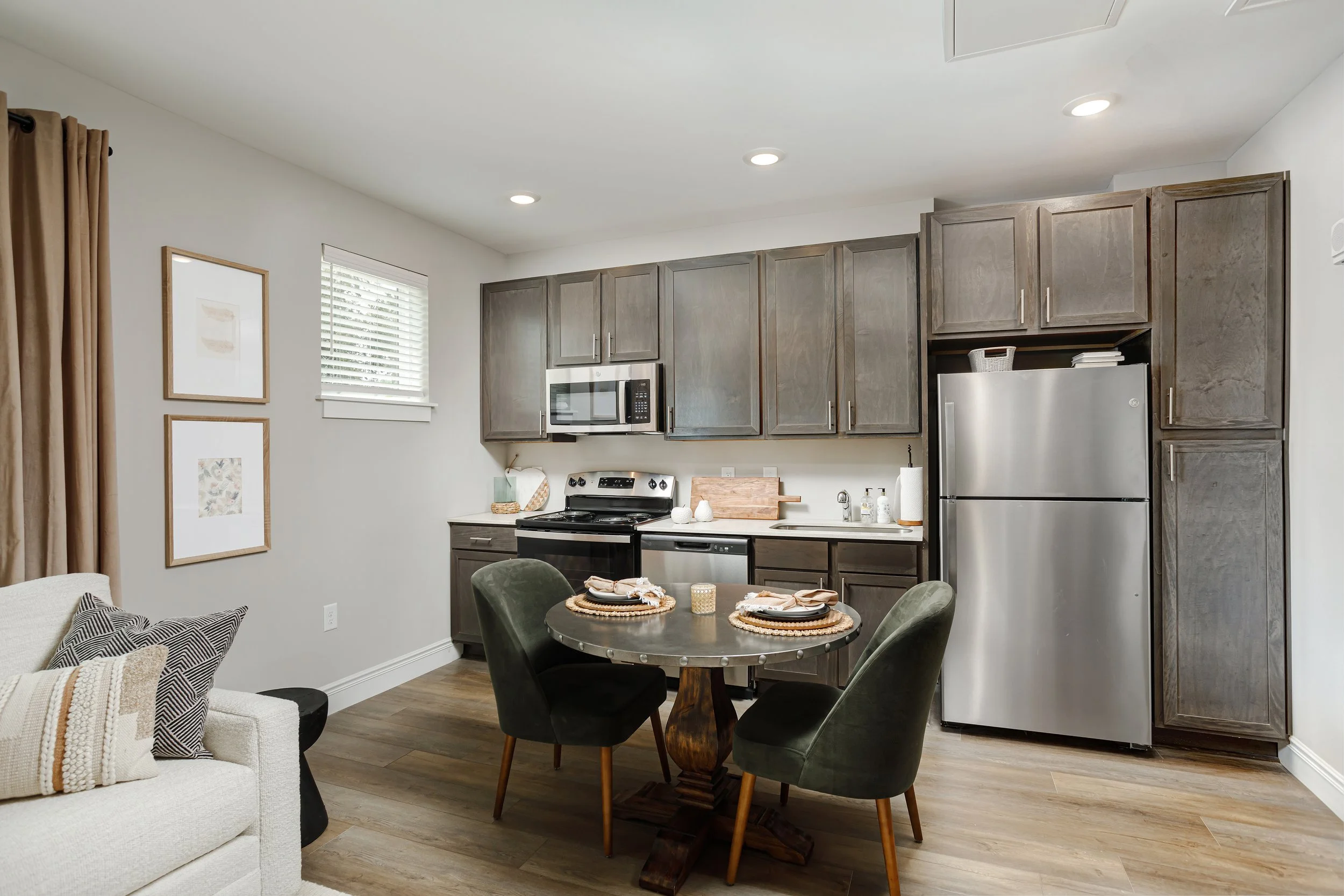 Modern kitchen and dining area with gray cabinets, stainless steel appliances, a round wooden dining table with four chairs, and decor including framed artwork and tableware.