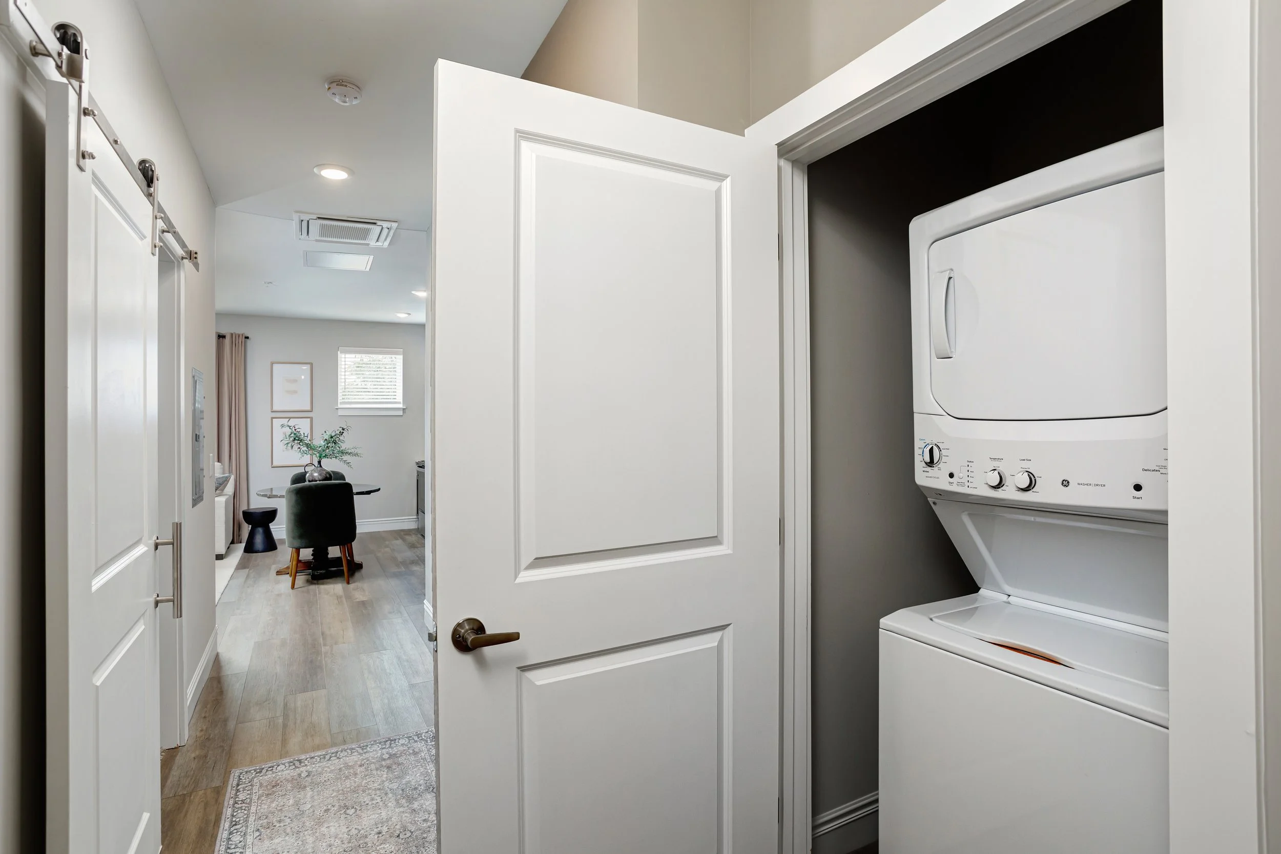 View inside a laundry closet with a stacked washer and dryer, located next to a doorway leading into a living area with a dining table and chairs.